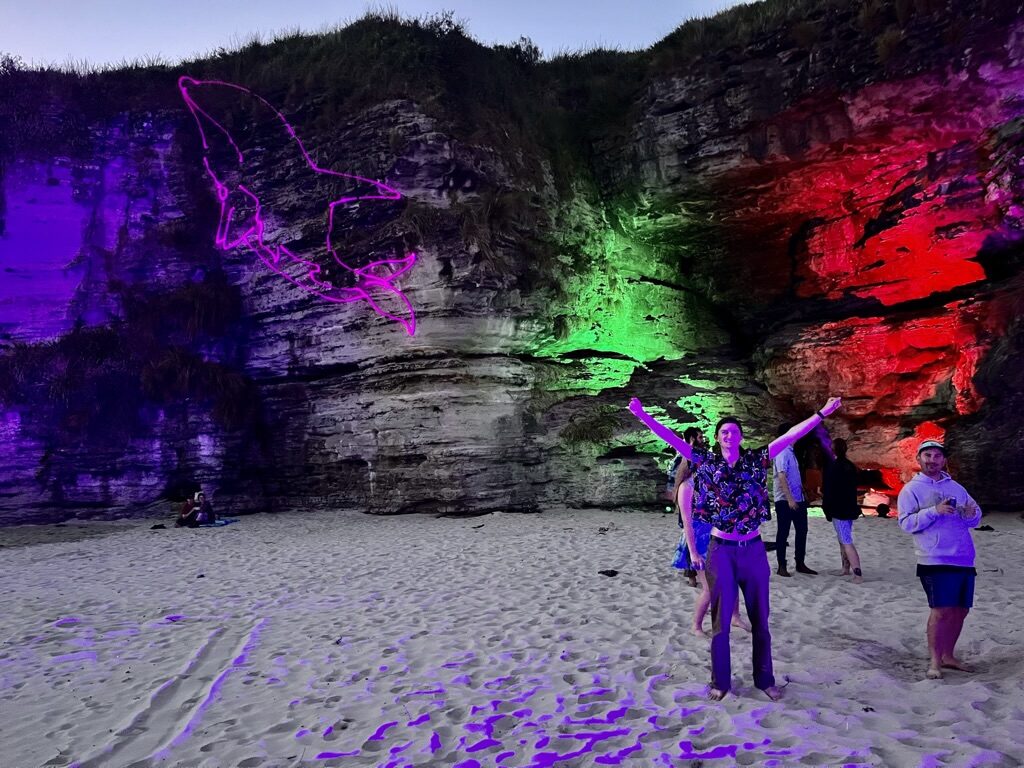 The cliff face at Cave Beach lit in purple, green and red at dusk, people dancing on the sand