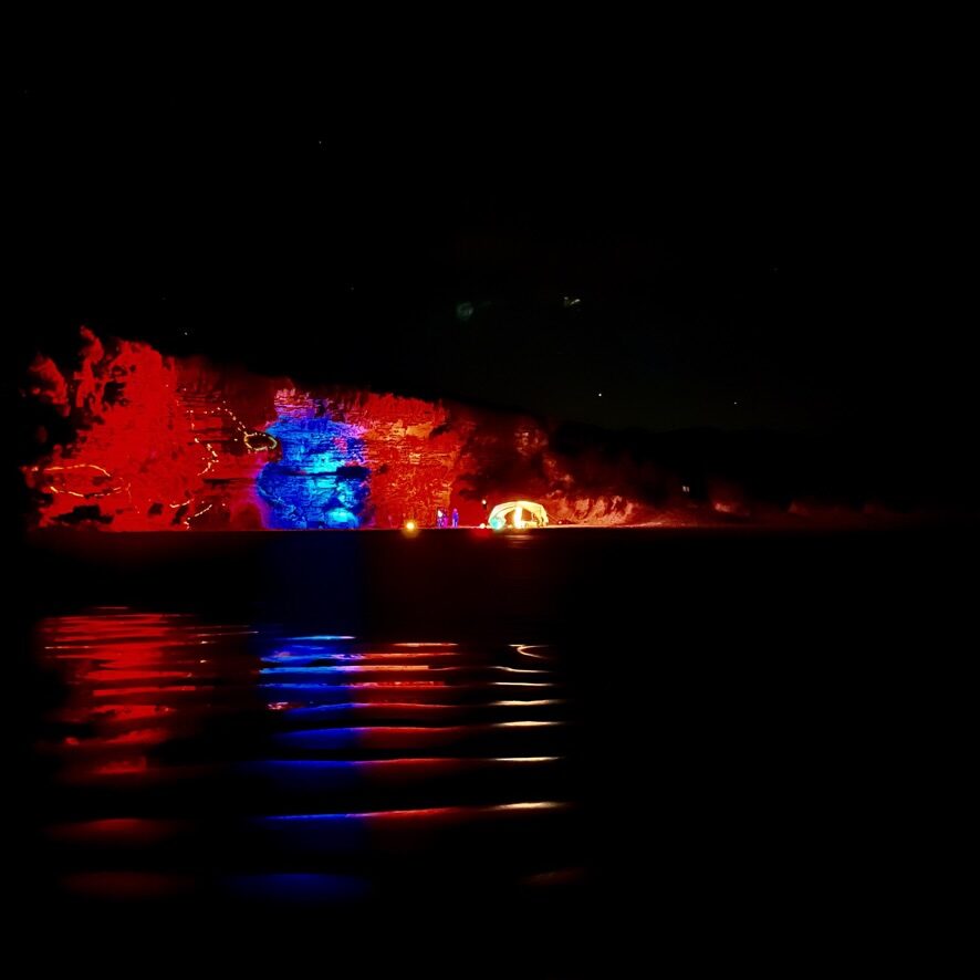 The same cliff at full dark, red and blue floodlights reflecting in long ribbons across the water